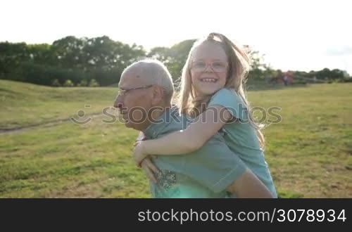 Adorable smiling girl in eyeglasses with long blonde hair enjoying piggyback ride with grandfather in park. Cheerful granddad carrying his granddaugter oh his back and running on green meadow in glow of sunset. Side view. Slow motion. Stabilized shot.
