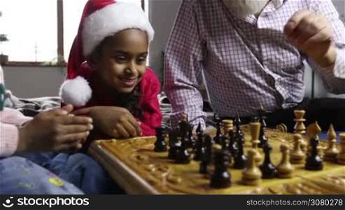 Adorable smiling mixed race little girl in santa hat lying on bed and watching chess match between caucasian grandpa and her younger sister on christmas eve. Hands of players moving chess pieces on chess board. Dolly shot. Slow motion.