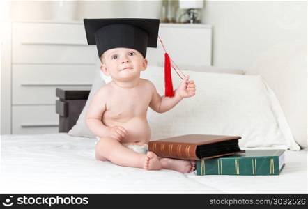 Adorable smiling toddler boy wearing mortarboard hat with books on bed. Smiling toddler boy wearing mortarboard hat with books on bed
