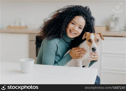 Adorable smiling woman embraces her favourite dog, expresses care and love, good attitiude to pet, poses against kitchen interior, drinks aromatic beverage. People, animals, relationship, affection
