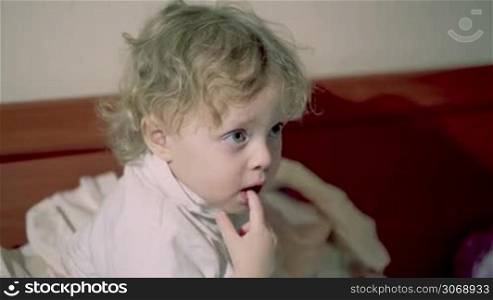 Adorable thoughtful little child with curly blond hair sitting chewing a finger and staring ahead with a pensive expression watching cartoons on tv