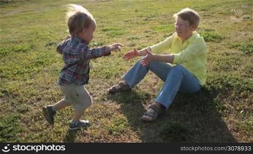 Adorable toddler grandson running into open arms of beloved grandmother in park while granny sitting on green grass. Attractive senior woman playing with child, lifting kid up while lying on her back on lawn and enjoying time together. Slow motion.