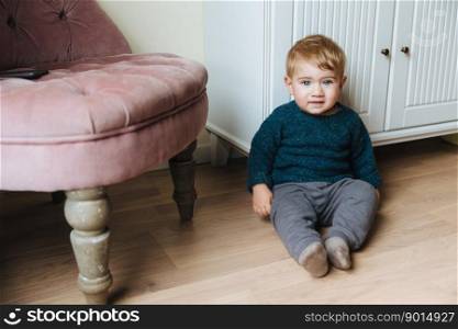 Adorable toddler with charming blue eyes and blonde hair, sits on floor against home interior, plays alone. Sweet baby boy looks directly innto camera, has pleasant appearance. Childhood concept