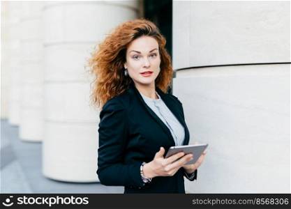 Adorable woman with curly hair wearing black suit and white blouse, holding tablet computer, typing messages or searching Internet while standing outdoor. Pretty businesswoman with modern device