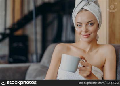 Adorable woman with natural makeup and healthy skin, poses bare shoulders, wrapped in bath towel, drinks tea or coffee, sits on comfortable sofa against home interior. People, hygiene, relaxation