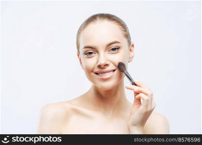 Adorable young brunette woman applies cosmetic tonal foundation with beauty brush, smiles tenderly, has healthy skin, well cared body, isolated on white background. Women, skin care, makeup concept