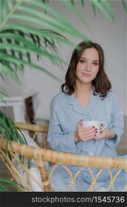 Adorable young female has charming look, dressed in striped domestic costume, drinks hot beverage, sits in wicker chair, enjoys rest and day off. People, lifestyle and recreation time concept
