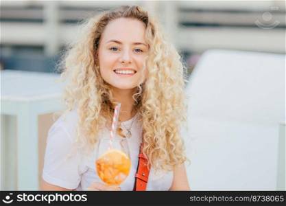 Adorable young female has toothy smile, fresh skin, blue eyes, drinks summer fresh cocktail, looks happily at camera, has unforgettable rest with friends during summer vacation. Leisure concept