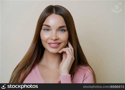 Adorable young female model smiles pleasantly, has long dark hair, looks directly at camera with satisfied expression, has perfect clean skin, wears rosy jumper, isolated over beige background