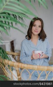 Adorable young woman in striped domestic costume, with a charming look, enjoys her day off, sipping a hot beverage in a wicker chair. People, lifestyle, and recreation time concept.