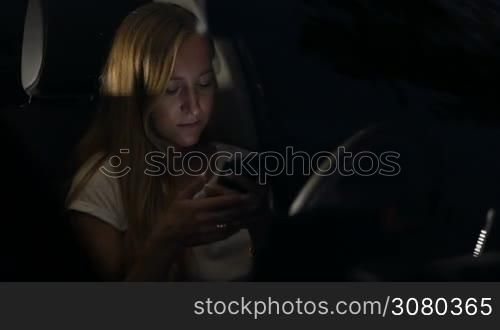 Adorable young woman with amazing long straight blonde hair sitting in car using mobile phone at night in the flashing lights of passing traffic. Attractive female driver surfing the net with smart phone on a summer night in her vehicle.