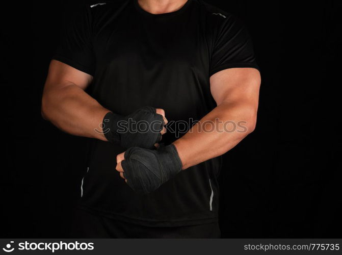 adult athlete in black uniform is standing in a rack with strained muscles, his hands are wrapped in a black textile bandage, dark background