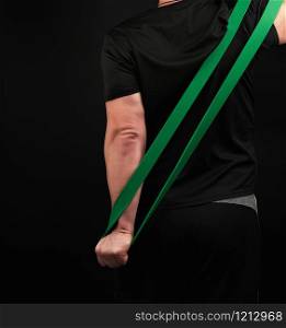 adult athlete in black uniform stands with his back and stretches a green sports elastic band, exercises for the muscles of the arms, dark background