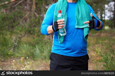 adult athlete is standing in the middle of nature in a blue uniform with a green towel, in one hand a transparent plastic water bottle with the other hand shows a gesture