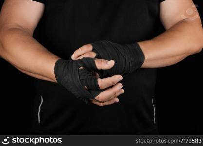 adult athlete stand in black clothes and wrap his hands in black textile elastic bandage before training, black background, muscular body