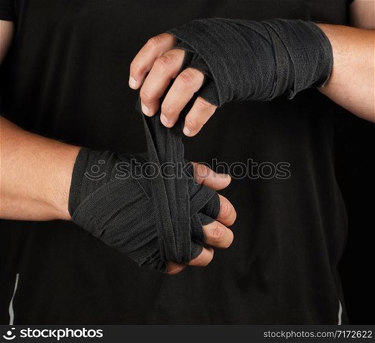 adult athlete stand in black clothes and wrap his hands in black textile elastic bandage before training, black background, muscular body