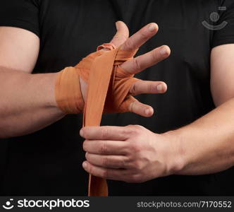 adult athlete stand in black clothes and wrap his hands in red textile elastic orange bandage before training, black background, muscular body