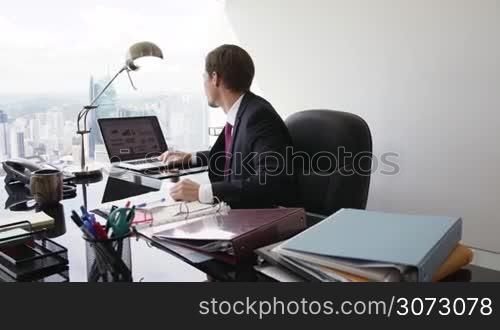 Adult businessman sitting in modern office with beautiful sight of the city. The man turns to the camera and smiles in portrait position. Medium shot, steadicam shot in slowmotion 60p