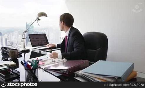 Adult businessman sitting in modern office with beautiful sight of the city. The man turns to the camera and smiles in portrait position. Medium shot, steadicam shot in slowmotion 60p