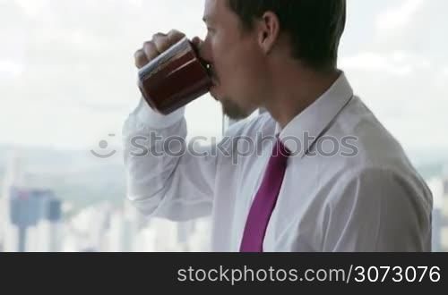 Adult businessman sitting on desk in modern office and reading news on tablet pc with a cup of coffee. The man looks out of the window and contemplates the city and skyscrapers. Medium shot