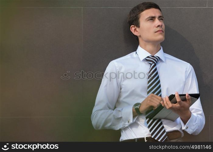 Adult businessman using tablet computer outdoors leaning on wall of office building