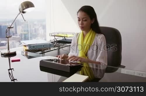 Adult businesswoman sitting in modern office with beautiful sight of the city. The secretary writes on tablet pc, then turns to the camera and smiles in portrait position. Medium shot, Slow motion, steadicam shot
