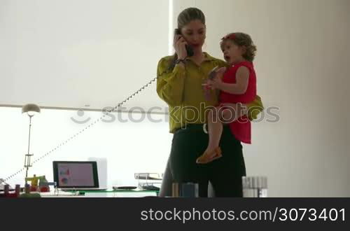 Adult businesswoman with little daughter in office. The multitasking mom holds her child and talks on telephone with client