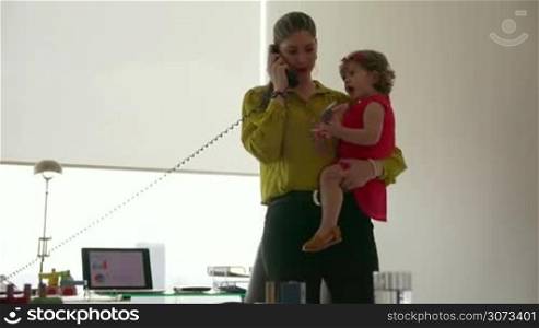 Adult businesswoman with little daughter in office. The multitasking mom holds her child and talks on telephone with client