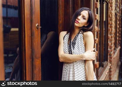 Adult caucasian woman standing by the wooden door building entrance looking to the camera sad holding her arm lonely frustrated leaning wearing dress in summer day love loneliness nostalgia concept