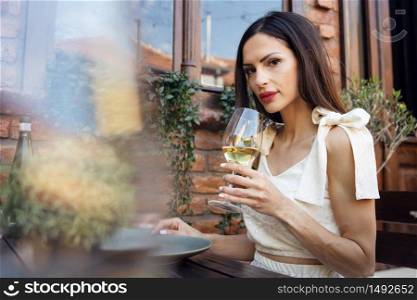 Adult caucasian woman wearing white dress sitting by the table in the backyard by the house or in cafe or restaurant having a glass of wine looking to the camera