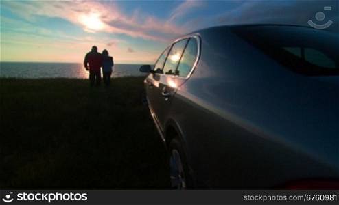 Adult couple silhouette near his car enjoying view of the sea coast at sunset