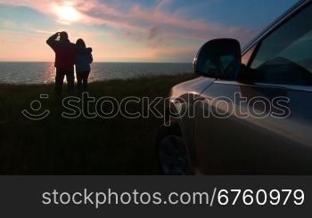 Adult couple silhouette near his car on sea coast watching the sunset