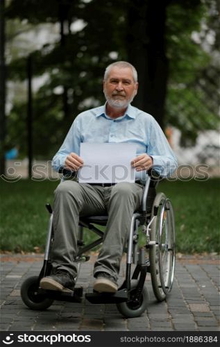 Adult disabled man in wheelchair holds empty paper sheet in summer park. Paralyzed people and disability, handicap overcoming. Alone handicapped male person leisures in public place. Adult disabled man in wheelchair holds paper sheet