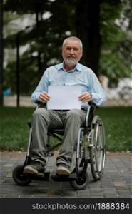 Adult disabled man in wheelchair holds empty paper sheet in summer park. Paralyzed people and disability, handicap overcoming. Alone handicapped male person leisures in public place. Adult disabled man in wheelchair holds paper sheet