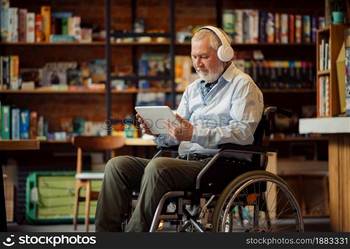 Adult disabled man in wheelchair listen to music in headphones, disability, cafeteria interior on background. Handicapped older male person, paralyzed people in public places. Adult disabled man in wheelchair listen to music