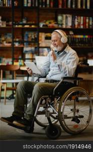 Adult disabled man in wheelchair listen to music in headphones, disability, cafeteria interior on background. Handicapped older male person, paralyzed people in public places. Adult disabled man in wheelchair listen to music