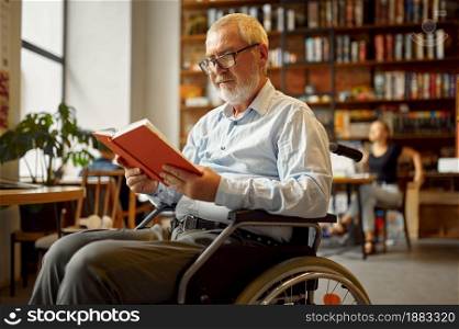Adult disabled man in wheelchair reading a book, disability, bookshelf and university library interior on background. Handicapped older male person, paralyzed people get knowledge. Adult disabled man in wheelchair reading a book