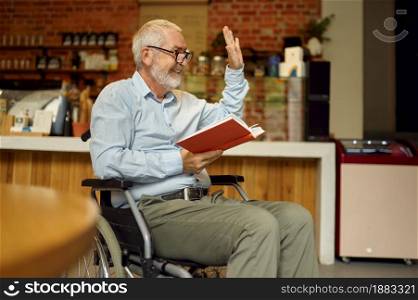 Adult disabled man in wheelchair reading a book, disability, bookshelf and university library interior on background. Handicapped older male person, paralyzed people. Adult disabled man in wheelchair reading a book