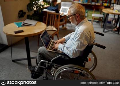 Adult disabled man in wheelchair using laptop, top view, disability, bookshelf and university library interior on background. Handicapped older male person, paralyzed people get knowledge. Adult disabled man using laptop, top view