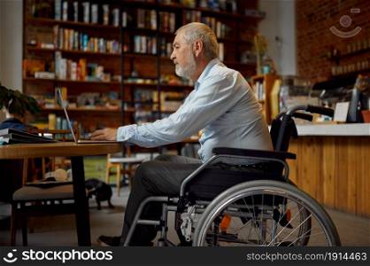 Adult disabled man in wheelchair using laptop, top view, disability, bookshelf and university library interior on background. Handicapped older male person, paralyzed people get knowledge. Adult disabled man using laptop, top view
