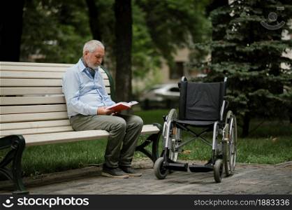 Adult disabled man with wheelchair sits on a bench in summer park. Paralyzed people and disability, handicap overcoming. Handicapped male person leisures in public place. Adult disabled man with wheelchair sits on a bench