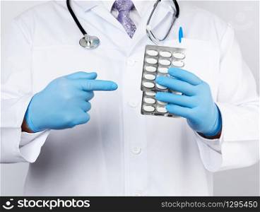 adult doctor therapist is dressed in a white uniform coat and blue sterile gloves is standing and holding a stack of pills in blister packs, white background
