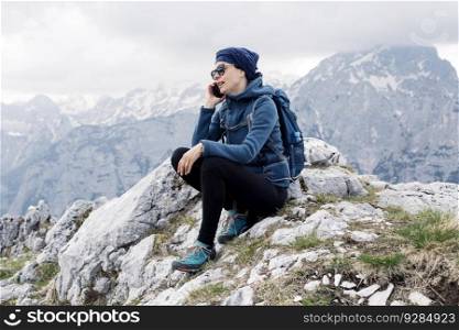 Adult female hiker in  blue jacket sitting on the peak of rock and talking on the phone