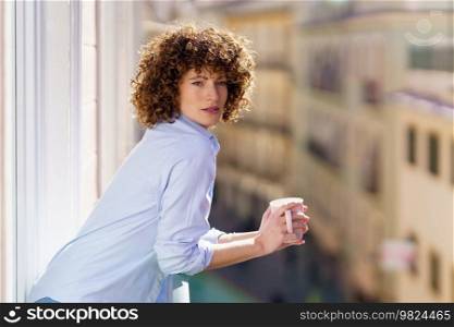 Adult female with brown curly hair leaning on railing of balcony while enjoying cup of hot beverage against blurred background. Curly haired woman drinking coffee on balcony