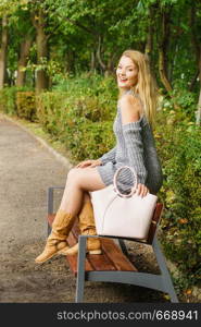 Adult feminine woman wearing long grey jumper sweater holding bag standing relaxing in park. Autumn fashion concept.. Fashion model relaxing in park