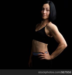 Adult girl with a sports figure in black bra and black shorts standing on a dark background, muscular body, black hair, low key