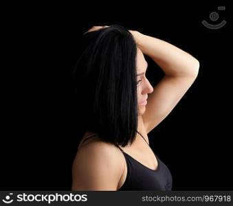 Adult girl with a sports figure in black bra standing on a dark background, model is turned sideways and straightens her black hair, low key
