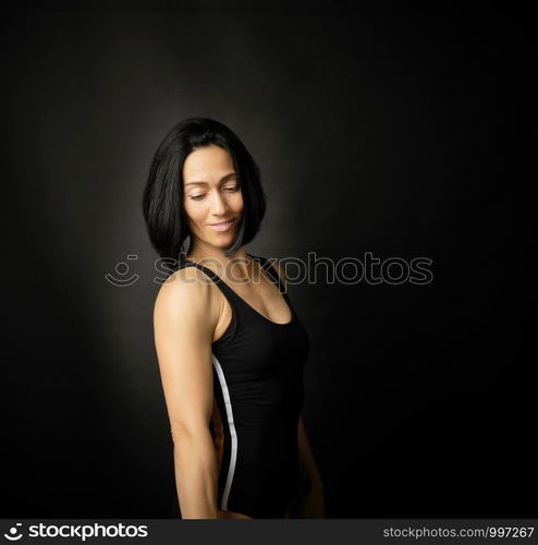 Adult girl with a sports figure standing on a dark background, muscular body, black hair, low key