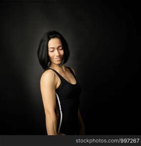 Adult girl with a sports figure standing on a dark background, muscular body, black hair, low key
