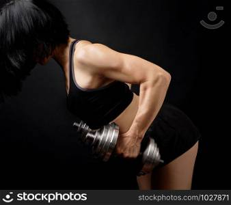 adult girl with black hair is dressed in a sports bra and short shorts is doing physical exercises on the arm muscles with a steel dial dumbbell, low key